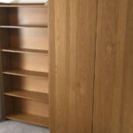 Property for sale by Auction A wooden wardrobe with closed doors stands next to an open bookcase with empty shelves. Both pieces of furniture are placed on a carpeted floor in a room with neutral walls. Presented by World of Auctions