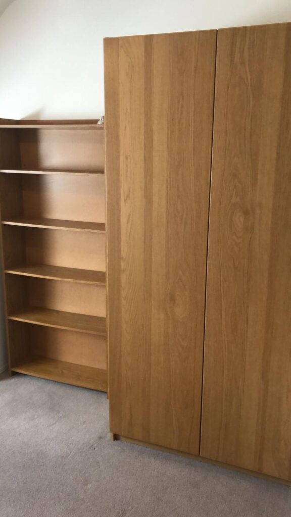 Property for sale by Auction A wooden wardrobe with closed doors stands next to an open bookcase with empty shelves. Both pieces of furniture are placed on a carpeted floor in a room with neutral walls. Presented by World of Auctions