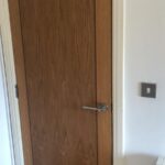 Property for sale by Auction A closed wooden door with a silver handle is shown against a white wall. To the right of the door, a light switch is mounted on the wall. The floor is carpeted in a neutral color. Presented by World of Auctions