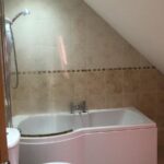 Property for sale by Auction A bathroom with a white toilet and a bathtub. The walls and floor are tiled in light beige. The bathtub is fitted into the corner under a sloped ceiling with a showerhead above it. The lighting is bright and modern. Presented by World of Auctions