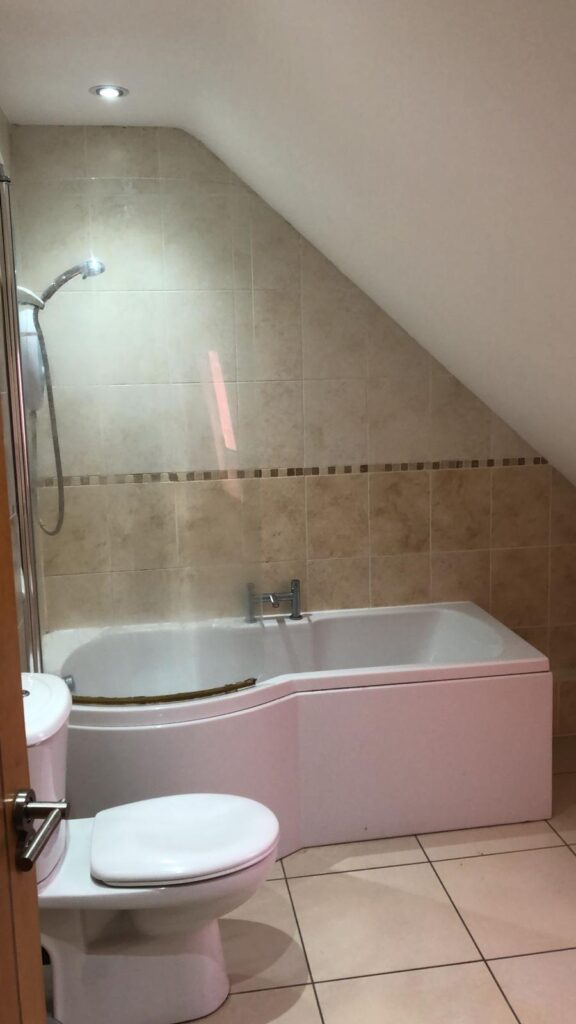 Property for sale by Auction A bathroom with a white toilet and a bathtub. The walls and floor are tiled in light beige. The bathtub is fitted into the corner under a sloped ceiling with a showerhead above it. The lighting is bright and modern. Presented by World of Auctions