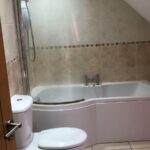 Property for sale by Auction A cozy bathroom with beige tiled walls and floor, featuring a white bathtub with a showerhead above it, and a toilet beside the tub. The ceiling is sloped, adding to the room's charm. A small trash bin is placed near the toilet. Presented by World of Auctions