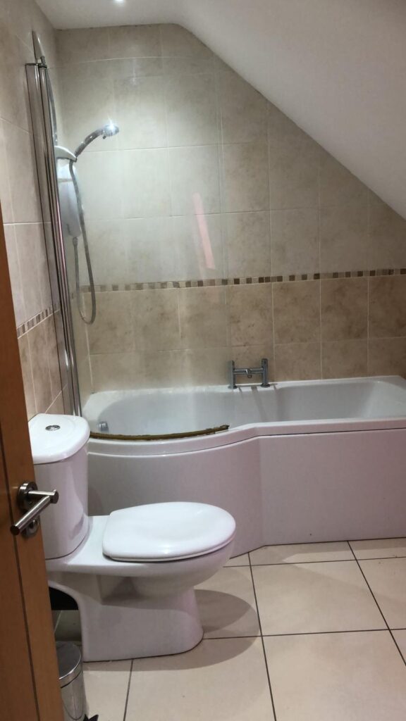 Property for sale by Auction A cozy bathroom with beige tiled walls and floor, featuring a white bathtub with a showerhead above it, and a toilet beside the tub. The ceiling is sloped, adding to the room's charm. A small trash bin is placed near the toilet. Presented by World of Auctions