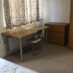 Property for sale by Auction A simple room with a wooden desk and chair beside a chest of drawers, under a window covered with leaf-patterned curtains. A bare mattress is visible in the foreground on a carpeted floor. Presented by World of Auctions