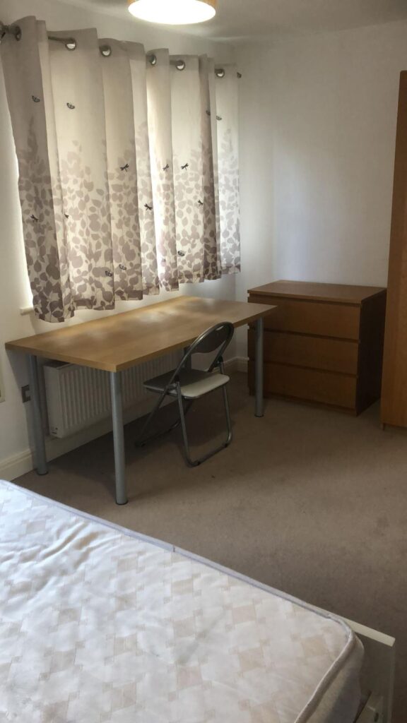 Property for sale by Auction A simple room with a wooden desk and chair beside a chest of drawers, under a window covered with leaf-patterned curtains. A bare mattress is visible in the foreground on a carpeted floor. Presented by World of Auctions