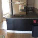 Property for sale by Auction A modern kitchen with wooden flooring features a large black countertop, a built-in oven, stovetop, and range hood. Light-colored cabinets line the back wall. A glass table is partially visible in the foreground. A fire extinguisher is on the counter. Presented by World of Auctions