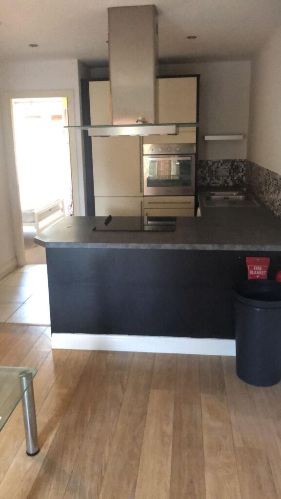 Property for sale by Auction A modern kitchen with wooden flooring features a large black countertop, a built-in oven, stovetop, and range hood. Light-colored cabinets line the back wall. A glass table is partially visible in the foreground. A fire extinguisher is on the counter. Presented by World of Auctions