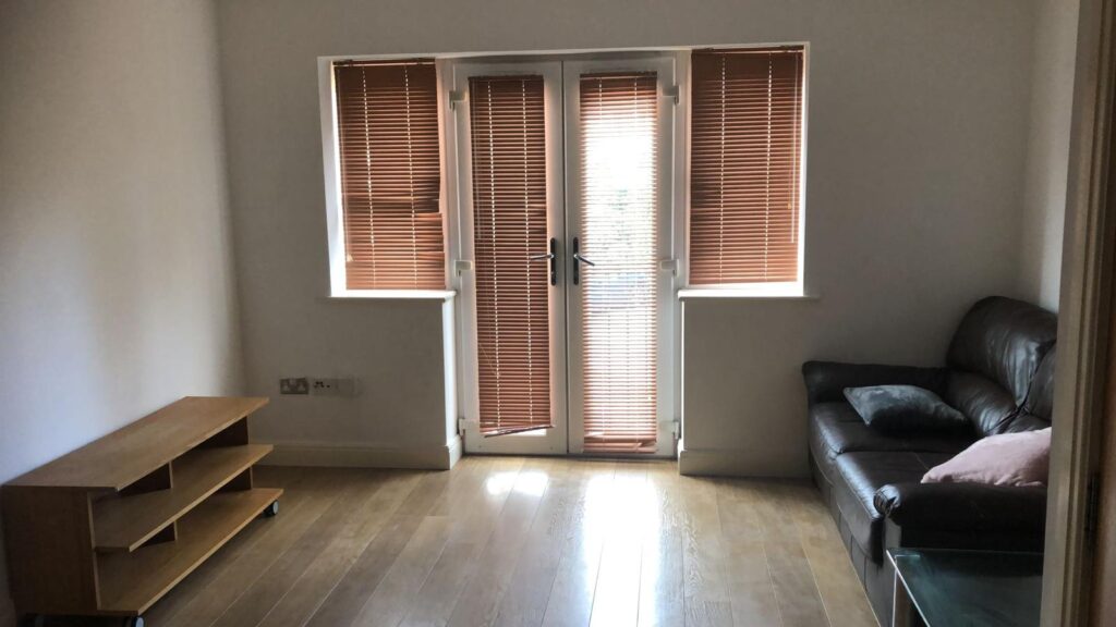 Property for sale by Auction A minimalist living room with wooden flooring, a brown leather sofa with pillows, a wooden TV stand, and large windows with brown blinds allowing filtered light to enter. Presented by World of Auctions