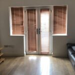 Property for sale by Auction A minimalist living room with wooden flooring, a brown leather sofa with pillows, a wooden TV stand, and large windows with brown blinds allowing filtered light to enter. Presented by World of Auctions