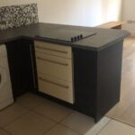 Property for sale by Auction A modern kitchen with a dark countertop and integrated stove. Below are three drawers with metallic handles. A washing machine is partially visible on the left. The floor is tiled, and a wall features a mosaic tile backsplash. Presented by World of Auctions