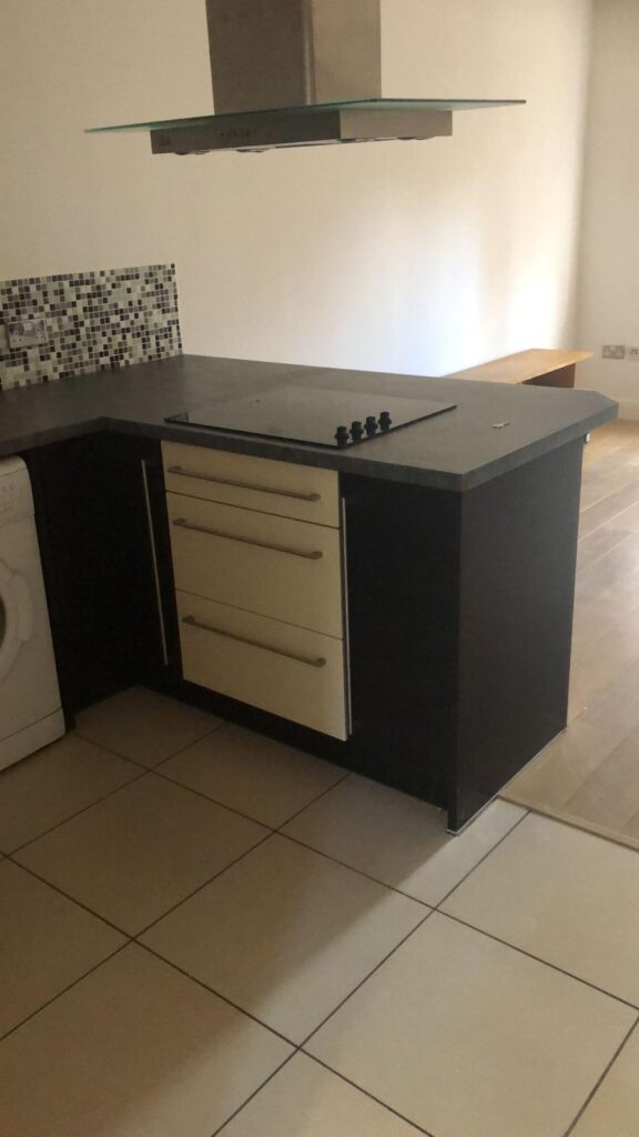 Property for sale by Auction A modern kitchen with a dark countertop and integrated stove. Below are three drawers with metallic handles. A washing machine is partially visible on the left. The floor is tiled, and a wall features a mosaic tile backsplash. Presented by World of Auctions