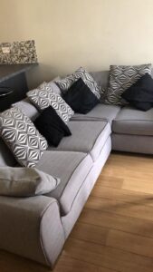 Property for sale by Auction A gray L-shaped couch with a variety of geometric-patterned and solid black cushions sits on a light wooden floor. A small section of a mosaic-tiled wall is visible in the background. Presented by World of Auctions