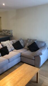 Property for sale by Auction A cozy living room with a light gray sectional sofa adorned with geometric-patterned and solid black throw pillows. A wooden coffee table sits in front, and a mosaic tile backsplash is partially visible in the background. Presented by World of Auctions