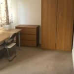 Property for sale by Auction A simple, unfurnished bedroom with a wooden wardrobe, a small chest of drawers, a desk, and a folding chair. There are floral curtains on the window, and a bed partially visible at the bottom of the image. Presented by World of Auctions