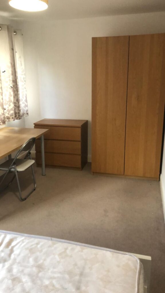 Property for sale by Auction A simple, unfurnished bedroom with a wooden wardrobe, a small chest of drawers, a desk, and a folding chair. There are floral curtains on the window, and a bed partially visible at the bottom of the image. Presented by World of Auctions
