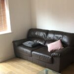 Property for sale by Auction A small living room with a brown leather sofa, a pink and a gray cushion. There's a window with wooden blinds, and a glass coffee table in the foreground on a light wooden floor. Walls are plain white. Presented by World of Auctions