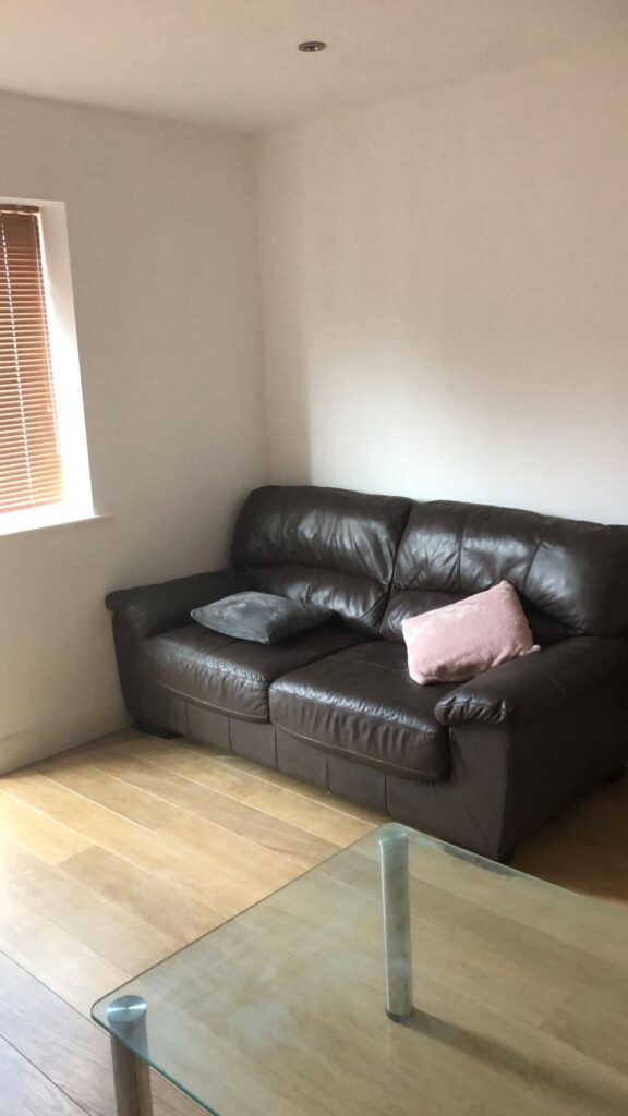 Property for sale by Auction A small living room with a brown leather sofa, a pink and a gray cushion. There's a window with wooden blinds, and a glass coffee table in the foreground on a light wooden floor. Walls are plain white. Presented by World of Auctions