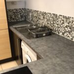 Property for sale by Auction A modern kitchen with a gray countertop and a mosaic-tiled backsplash. It features a stainless steel sink with a chrome faucet, a dish rack, an induction cooktop, and a washing machine beneath the counter. Light wooden cabinets are visible. Presented by World of Auctions