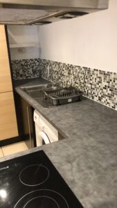 Property for sale by Auction A modern kitchen with a gray countertop and a mosaic-tiled backsplash. It features a stainless steel sink with a chrome faucet, a dish rack, an induction cooktop, and a washing machine beneath the counter. Light wooden cabinets are visible. Presented by World of Auctions