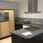 Property for sale by Auction A modern kitchen with light wood and dark countertops, featuring a built-in oven, an electric stovetop on an island with a stainless steel vent hood, and a mosaic tile backsplash. There's a small white appliance and a paper on the counter. Presented by World of Auctions