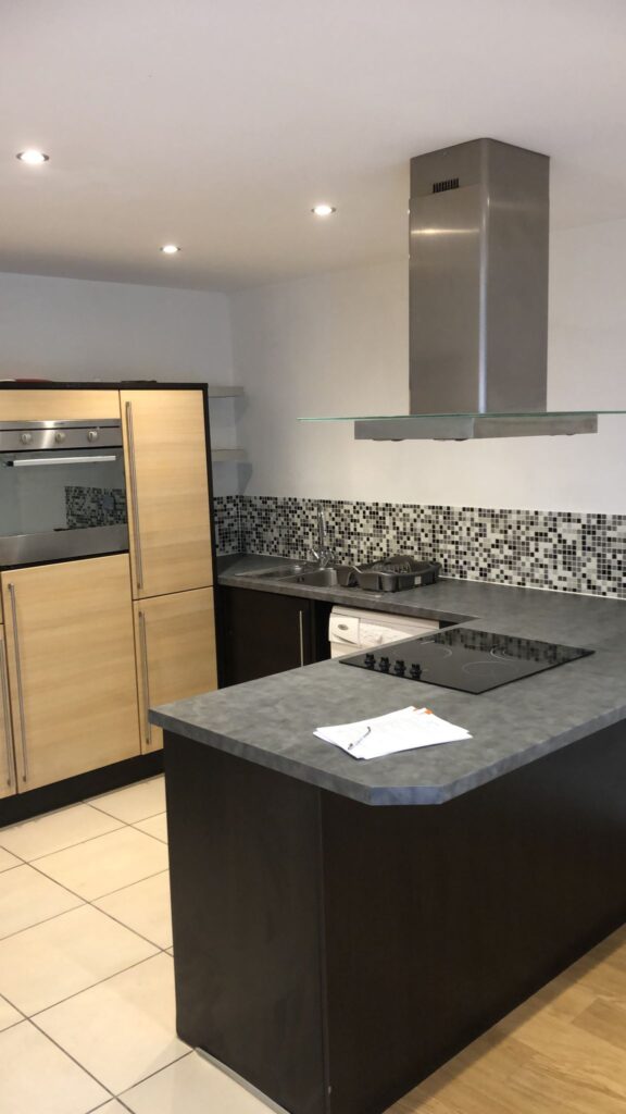 Property for sale by Auction A modern kitchen with light wood and dark countertops, featuring a built-in oven, an electric stovetop on an island with a stainless steel vent hood, and a mosaic tile backsplash. There's a small white appliance and a paper on the counter. Presented by World of Auctions