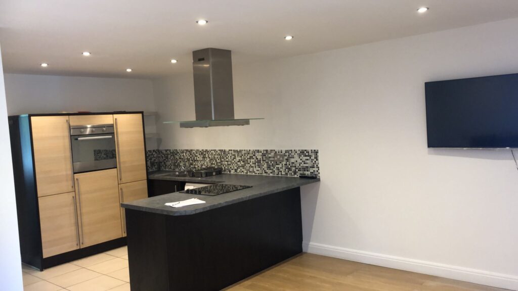Property for sale by Auction A modern kitchen with light wood cabinets, a built-in oven, a stovetop with an overhead range hood, and a black countertop. A flat-screen TV is mounted on the white wall to the right. The backsplash features gray and black mosaic tiles. Presented by World of Auctions