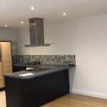 Property for sale by Auction A modern kitchen with light wood cabinets, a built-in oven, a stovetop with an overhead range hood, and a black countertop. A flat-screen TV is mounted on the white wall to the right. The backsplash features gray and black mosaic tiles. Presented by World of Auctions