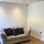 Property for sale by Auction A small living room with a beige sofa adorned with dark throw pillows, placed against a plain white wall. The room has hardwood floors and is softly lit by recessed ceiling lights, with a black door visible on the left. Presented by World of Auctions