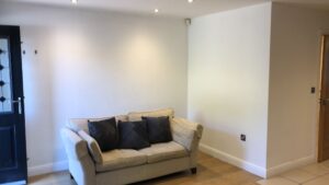 Property for sale by Auction A small living room with a beige sofa adorned with dark throw pillows, placed against a plain white wall. The room has hardwood floors and is softly lit by recessed ceiling lights, with a black door visible on the left. Presented by World of Auctions