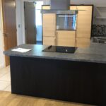 Property for sale by Auction A modern kitchen with light wooden cabinets, a dark kitchen island, and a black electric stovetop. A metallic range hood is above the island. The floor is a mix of wood and tile. Bright lighting illuminates the space. Presented by World of Auctions