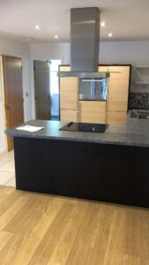 Property for sale by Auction A modern kitchen with light wooden cabinets, a dark kitchen island, and a black electric stovetop. A metallic range hood is above the island. The floor is a mix of wood and tile. Bright lighting illuminates the space. Presented by World of Auctions