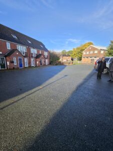 Property for sale by Auction A paved courtyard is bordered by two brick buildings under a clear blue sky. Three people stand near a white vehicle parked on the right. In the background, there are trees and additional buildings. Presented by World of Auctions