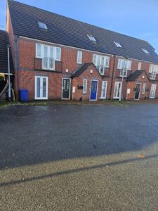 Property for sale by Auction A row of modern, three-story red-brick townhouses with dark roofs. Each has a blue, green, or black front door and small windows on the top floor. A wet pavement and a blue recycling bin are visible in front of the houses. Presented by World of Auctions