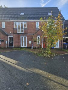 Property for sale by Auction A two-story brick house with a slate roof, featuring multiple doors and windows. A small tree with autumn leaves stands in front. The sky is clear, and shadows stretch across the driveway. Bicycles are parked near the entrance. Presented by World of Auctions
