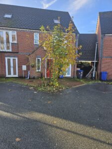 Property for sale by Auction A two-story brick house with dark roof tiles and a small tree with autumn leaves in front. Recycling bins are positioned near the garage on the right. The driveway and road are visible in the foreground under a clear blue sky. Presented by World of Auctions