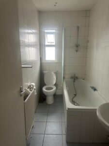 Property for sale by Auction Nestled at 99 Lower Addiscombe Road, this small apartment bathroom boasts white tiled walls and floor. It features a bathtub with a handheld showerhead, a toilet, and a sink. A window allows natural light to grace the space, complementing the practical radiator stocked with toilet paper rolls. Presented by World of Auctions