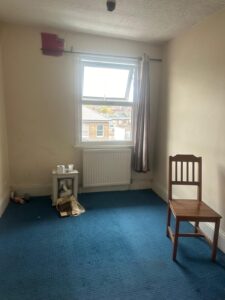 Property for sale by Auction A small, sparsely furnished room in an apartment at 99 Lower Addiscombe Road features a blue carpet. There's a wooden chair, a small table with scattered papers, toilet rolls, and a soft toy. A single window lets in natural light. The walls are plain, and a curtain hangs partly open. Presented by World of Auctions