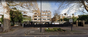 Property for sale by Auction Street view of a Maida Vale residential area in London, featuring brown brick apartments behind a black fence. Large trees with bare branches line the street, and a small roundabout with signs and a lamp post is in the foreground—a potential hotel redevelopment opportunity awaits. Presented by World of Auctions