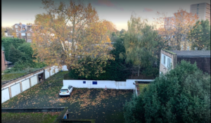 Property for sale by Auction Aerial view of a small parking lot surrounded by trees with autumn leaves. A single white car is parked near a row of garages, hinting at Maida Vale London’s serene charm. Residential buildings and a city skyline in the background suggest exciting hotel redevelopment opportunities. Presented by World of Auctions