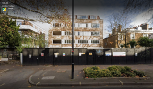 Property for sale by Auction Street view of a residential building in Maida Vale, London. The four-story structure is partially obscured by leafless trees and a construction fence, hinting at a hotel redevelopment opportunity. A lamppost and small garden area are visible in the foreground. Presented by World of Auctions