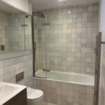 Property for sale by Auction This Wimbledon new home for sale boasts a modern bathroom with a glass shower over a bathtub, tiled walls, and a light wooden floor. The well-lit space features recessed lighting, a sink with cabinet, wall-mounted toilet, and a skylight. Presented by World of Auctions