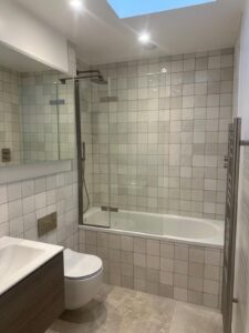 Property for sale by Auction This Wimbledon new home for sale boasts a modern bathroom with a glass shower over a bathtub, tiled walls, and a light wooden floor. The well-lit space features recessed lighting, a sink with cabinet, wall-mounted toilet, and a skylight. Presented by World of Auctions