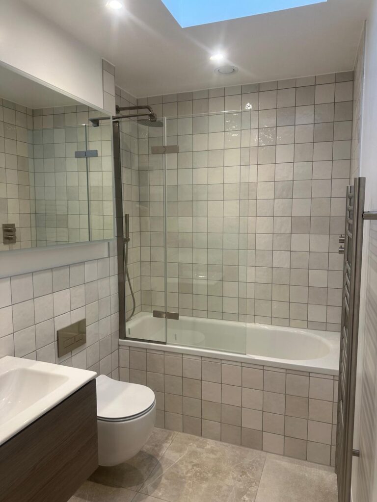 Property for sale by Auction This Wimbledon new home for sale boasts a modern bathroom with a glass shower over a bathtub, tiled walls, and a light wooden floor. The well-lit space features recessed lighting, a sink with cabinet, wall-mounted toilet, and a skylight. Presented by World of Auctions