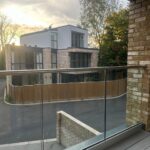 Property for sale by Auction View from a balcony overlooking a modern residential area at sunset. A glass railing in the foreground frames two contemporary houses with large windows and brick exteriors, nestled among trees—a perfect new home for sale in Wimbledon. Presented by World of Auctions