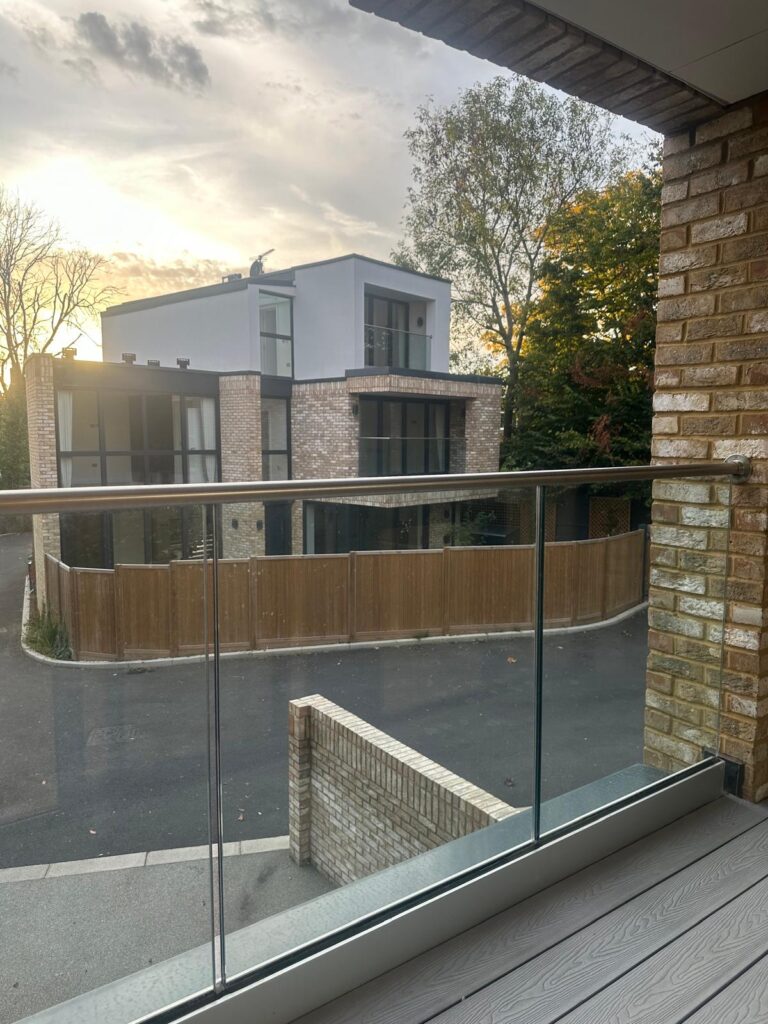 Property for sale by Auction View from a balcony overlooking a modern residential area at sunset. A glass railing in the foreground frames two contemporary houses with large windows and brick exteriors, nestled among trees—a perfect new home for sale in Wimbledon. Presented by World of Auctions