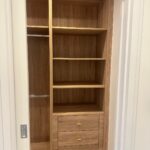 Property for sale by Auction A wooden closet with open shelves on the right, a metal rod for hanging clothes on the left, and three drawers with gold handles at the bottom. The closet features a natural wood finish and is framed by white walls—a perfect addition to your new Wimbledon home for sale. Presented by World of Auctions