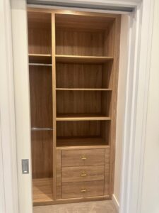 Property for sale by Auction A wooden closet with open shelves on the right, a metal rod for hanging clothes on the left, and three drawers with gold handles at the bottom. The closet features a natural wood finish and is framed by white walls—a perfect addition to your new Wimbledon home for sale. Presented by World of Auctions