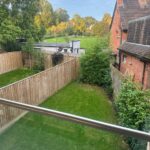 Property for sale by Auction View from a balcony overlooking a fenced yard with lush green grass and a few bushes. Adjacent to the yard is a red brick house with chimneys, reminiscent of Wimbledon charm. In the background, more trees and greenery stretch under a cloudy sky, hinting at your new home for sale. Presented by World of Auctions