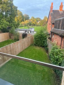 Property for sale by Auction View from a balcony overlooking a fenced yard with lush green grass and a few bushes. Adjacent to the yard is a red brick house with chimneys, reminiscent of Wimbledon charm. In the background, more trees and greenery stretch under a cloudy sky, hinting at your new home for sale. Presented by World of Auctions