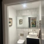 Property for sale by Auction This new home for sale features a modern bathroom with white tiled walls, a large mirror, and a rectangular sink on a sleek black vanity. A towel hangs from a rail beside it. A wall-mounted toilet sits beneath a framed picture, complementing the dark-colored floor—a Wimbledon gem indeed. Presented by World of Auctions